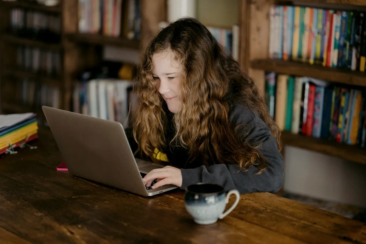 Teenage student smiling while working on her laptop in front of a bookshelf